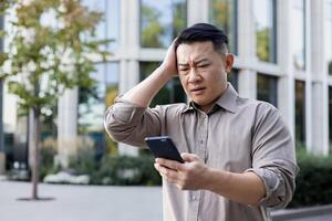 Upset young asian man standing near office building and holding head, reading bad news and message on phone. photo