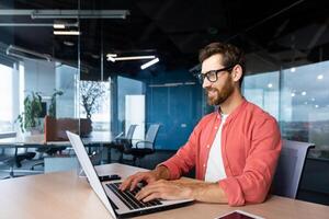 exitoso sonriente hombre trabajando dentro oficina con computadora portátil, empresario en rojo camisa sonriente y mecanografía en teclado en anteojos, programador trabajando software para programa. foto