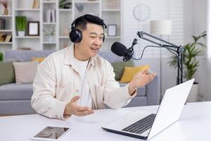 Happy Asian male podcaster recording audio content at home using laptop and studio microphone, engaging with online audience. photo