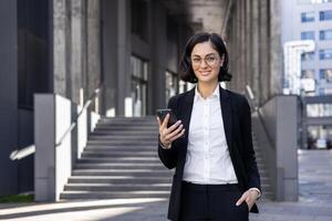 confidente hembra ejecutivo en un negocio traje sonriente y mensajes de texto en su móvil teléfono en frente de un moderno edificio. foto