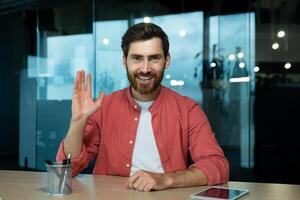 call, joyful smiling man waving hand greeting gesture working in evening time inside office, web camera view, online meeting with colleagues partners remotely. photo