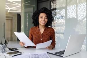 retrato de exitoso satisfecho y confidente mujer jefe financiero a lugar de trabajo, africano americano sonriente y mirando a cámara, mujer de negocios detrás papeleo dentro oficina trabajando con documentos. foto