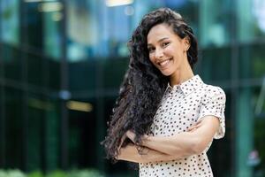 A cheerful Hispanic woman with curly long hair smiles brightly standing against a modern glass building backdrop. She is wearing a polka dot shirt. photo