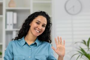 sonriente mujer en un casual camisa ondulación Hola durante un llamar, Proporcionar un atractivo cámara web ver pov en un hogar ajuste. foto