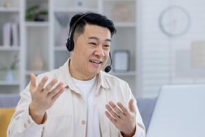 A cheerful Asian man engages in a lively conversation using a headset in a bright, neatly organized home office setting, demonstrating remote work communication. photo