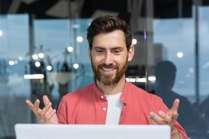 llamada en línea comunicación remoto, hombre sonriente y contento chateando con colegas en línea reunión, empresario mirando a ordenador portátil cámara web trabajando dentro moderno oficina edificio foto