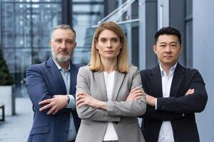 Successful and serious diverse team of three business people, man and woman focused looking at camera with arms crossed, portrait of co-workers outside office building photo