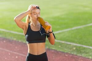 un ajuste joven mujer con auriculares es tomando un descanso a hidratar con un agua botella en un soleado atlético pista. foto