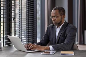 Serious focused male businessman working inside office with laptop, mature experienced african american man typing on keyboard, thinking reading from monitor. photo