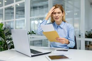 Shocked woman in formalwear touching forehead with arm while reading letter from open envelope in working space. Overworked manager receiving postal mail with warning from bank about debt payment. photo