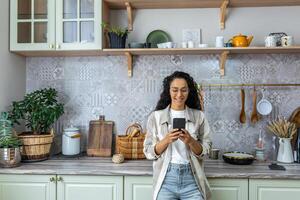 Hispano mujer usos móvil teléfono a hogar en el cocina, mujer sonrisas y se regocija, lee mensajes, navega web páginas, en línea comunicación con amigos y tener una cita. foto