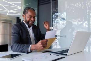 Senior african american man in suit sitting in office at desk and happy. Shows a victory gesture with his hand. He reads the letter in the envelope. Got a job, position, promotion. photo