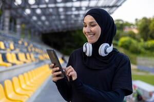 musulmán deportista en hijab a estadio con auriculares y teléfono en manos, mujer durante activo ejercicio y aptitud física, al aire libre escuchando a en línea música desde audio libros y podcasts aplicación foto