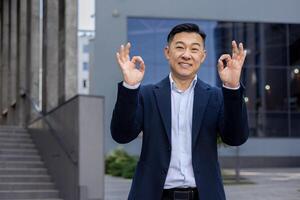 Confident male Asian executive showing 'OK' sign with both hands, expressing approval in an urban setting. photo