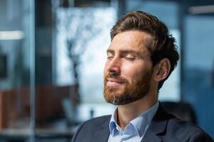 de cerca foto. retrato de un hermoso joven hombre quien cerrado su ojos y descansa a el lugar de trabajo. sentado relajado en el oficina. foto