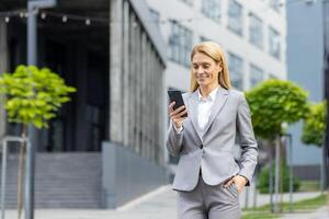 exitoso mujer jefe camina fuera de oficina edificio al aire libre, en negocio traje, mujer de negocios usos aplicación en teléfono, sonrisas contentamente lee Internet páginas, navega noticias, escribe texto mensaje, llamar. foto