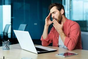 bostezando y aburrido hombre dentro oficina, sobrecargado de trabajo y demasiado cansado empresario con diario rutina trabajar, trabajador trabajando tarde con computadora portátil, preparando reporte en línea. foto