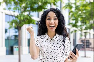 Joyful Latin American businesswoman with curly hair using smartphone, celebrating success outside office building. photo