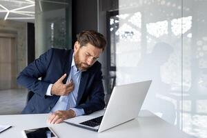 joven masculino empresario sufrimiento desde dolor y corazón ataque a lugar de trabajo. él se sienta a el escritorio en el oficina y sostiene su manos a su pecho. foto