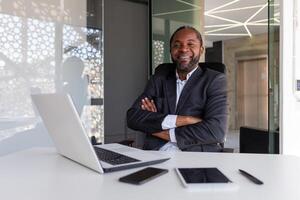 retrato de exitoso maduro adulto empresario, hombre con cruzado brazos sonriente y mirando a cámara, africano americano jefe financiero sentado a mesa con ordenador portátil. foto