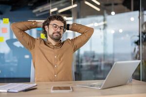 un joven profesional toma un momento a relajarse, reclinable con manos detrás cabeza a su espacio de trabajo con un ordenador portátil y tableta en frente de a él. foto