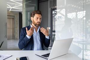 A serious young man works in the office, sits at a desk with a laptop and waves his hands in his face in the air from the heat, he does not feel well. photo