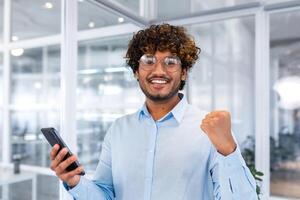 joven Hispano masculino alumno, persona de libre dedicación vistiendo lentes en pie en oficina y utilizando teléfono. él mira a el cámara, marca, se regocija, celebra, muestra un victoria gesto con su mano. foto