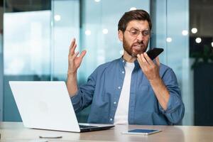 Angry businessman shouting on the phone, man gossiping with colleagues quarreling, inside a modern office building with a laptop photo