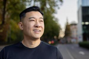 Close-up photo of a young Asian male athlete standing on a city street and smilingly looking to the side.