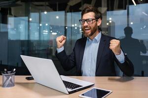 Happy and successful mature boss celebrating victory, senior businessman with beard holding hands up gesture of triumph and achievement, man in glasses and business suit working inside modern office. photo