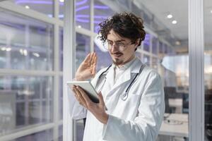 A doctor wearing a white lab coat and stethoscope holds a tablet inside a medical office of a clinic. Man talking call photo