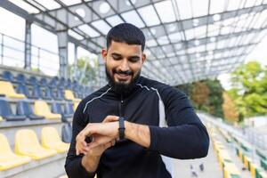 un sonriente, barbado hombre en ropa de deporte comprobación su aptitud rastreador después un rutina de ejercicio a un al aire libre estadio con asientos en el antecedentes. foto