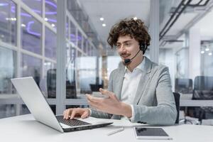 confidente joven adulto empresario con Rizado pelo utilizando ordenador portátil y auriculares en un oficina configuración, atractivo en corporativo trabajar. foto