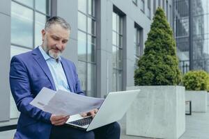 Senior handsome successful male engineer architect in suit works with laptop, documents, plan, drawings. Sitting on a bench near the office center, holding papers, looking around, smiling. photo