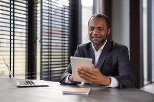 sonriente africano americano empresario trabajando en moderno oficina con tableta y computadora portátil, representando profesionalismo y éxito. foto