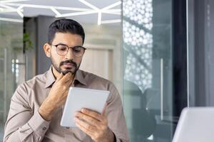 Contemplative Indian businessman in eyeglasses using a digital tablet, engrossed in deep thought at his modern office. photo