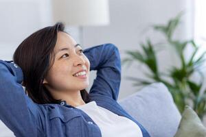 Closeup photo of a young beautiful Asian at home in the living room, woman has her hands behind her head and is resting, breathing fresh air, smiling and looking window, dreaming happy future