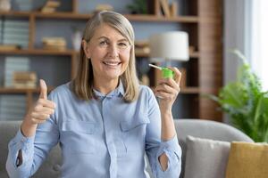Portrait of optimistic mature woman showing thumb up gesture while holding spray for throat while sitting on sofa. Smiling lady feeling relief after sprinkling cure in mouth for softening sore spot. photo
