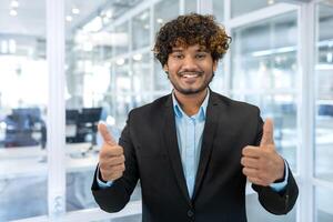 Portrait of successful hispanic businessman boss, manager in business suit and beard looking at camera and smiling standing near window, showing thumbs up, sign of success and achieving goals. photo