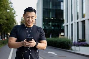 Smiling young Asian man doing morning jog with phone and headphones. Turns on music, podcast. photo