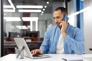 A focused professional man engaging in a phone conversation while working on a laptop in a modern office environment. photo