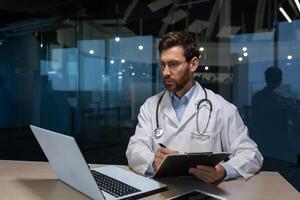 Mature doctor working on paper work inside a shabby office building, man in medical coat using laptop filling out medical documents concentrating and thinking photo