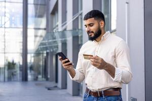 Confident young adult male using a smartphone and holding a credit card on a sunny day outside modern office buildings. Concept of mobile banking, e-commerce and modern lifestyle. photo