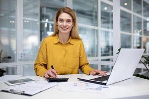 sonriente hembra jefe trabajando en brillante oficina a mesa en frente de computadora portátil, contando en calculadora, contento rubia sonriente y mirando a cámara. foto