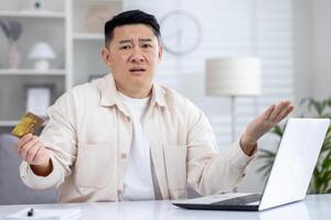 confuso asiático hombre sentado en ligero oficina a mesa en frente de computadora portátil, participación tarjeta en mano, trastornado acerca de ha fallado en línea compras, roto computadora, ha fallado en línea transacción. foto