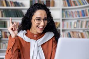 Idea concept. Excited young Indian woman wearing glasses sitting at a desk using a laptop and raising her index finger up in a classroom. Amazed, shocked lady, seeing, aha moment. photo