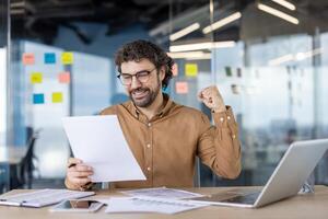 Excited male professional cheers with a fist pump while reviewing documents at his office desk, expressing success and positivity. photo