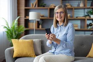maduro adulto mujer cómodamente sentado en un sofá, hojeada su teléfono inteligente en un moderno vivo habitación ajuste. foto