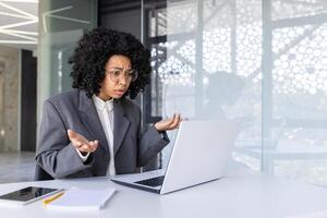 africano americano joven negocio mujer hablando en el oficina en un llamada en un computadora portátil, lanzamiento arriba su manos en frustración y decepción. foto