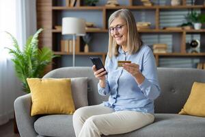 Cheerful mature woman sitting on the sofa, using her smartphone and holding a credit card, smiling as she shops online in a cozy living room setting. photo